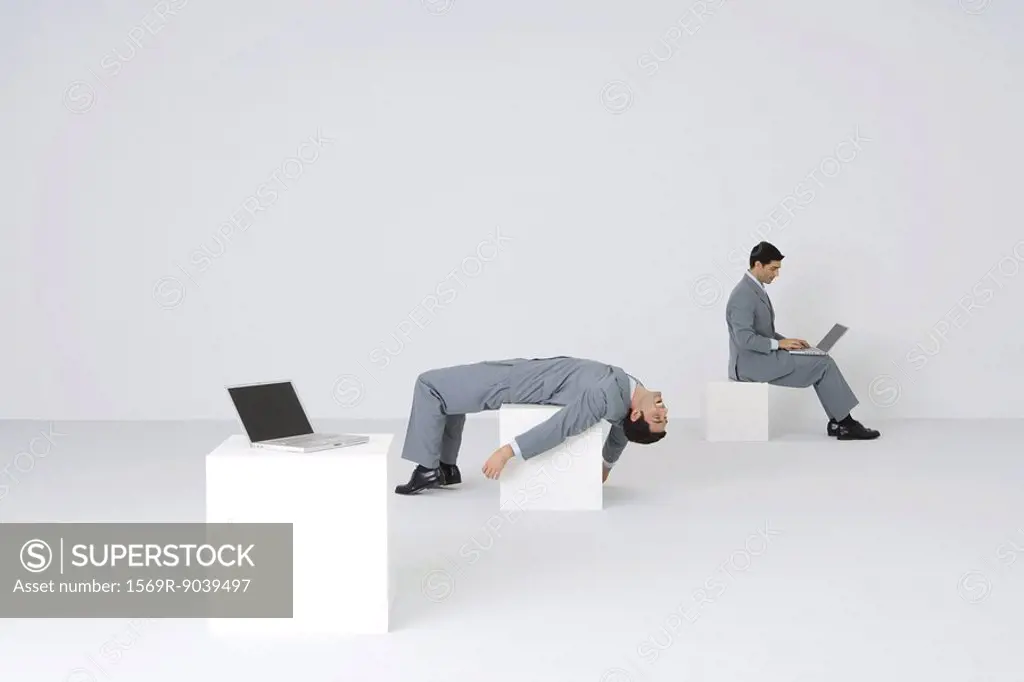 Identical businessmen, one using laptop while the other lies on back, exhausted