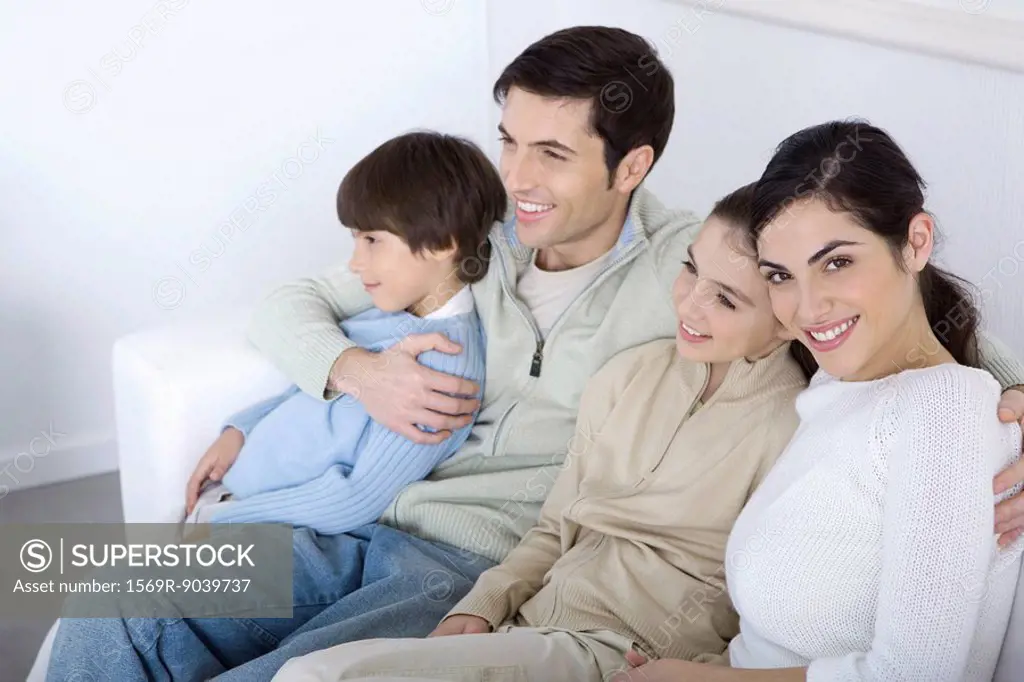 Family sitting together on sofa, looking away, woman smiling at camera