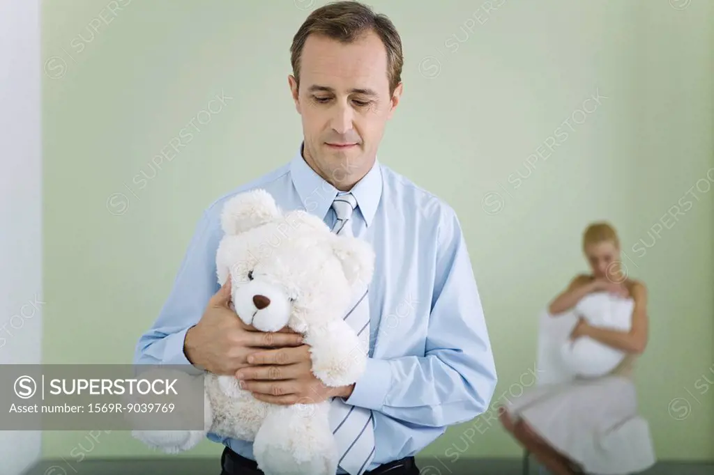 New father holding teddy bear, looking down, wife holding baby in background