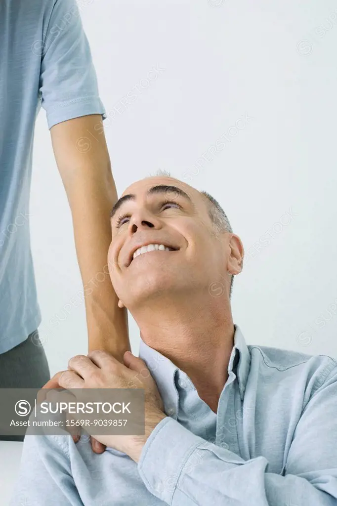 Mature man holding son´s hand on shoulder, smiling up over shoulder, cropped view