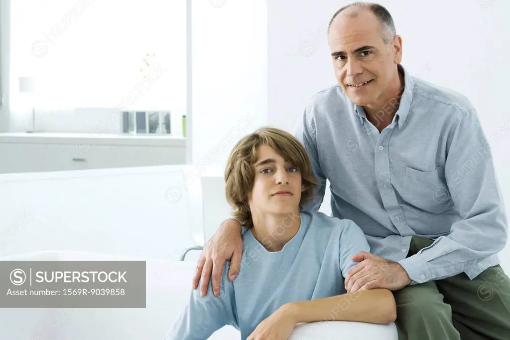 Father and teen son sitting together on sofa, looking at camera, portrait
