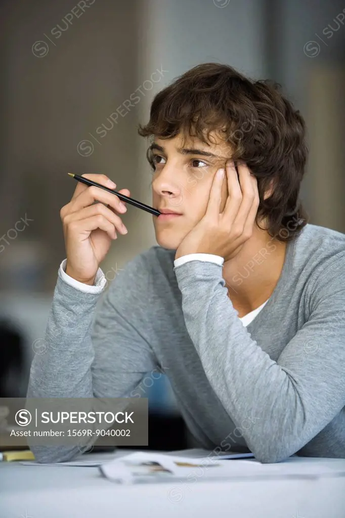 Young man with pen in mouth leaning on elbow, idly looking away