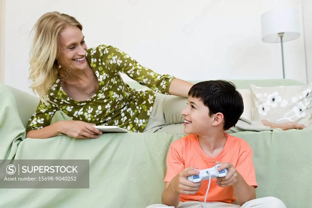 Young boy sitting on floor playing video game looking up at mother relaxing on sofa