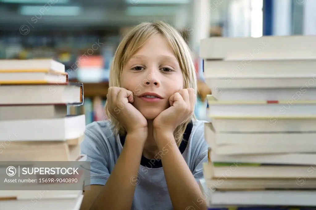 Boy leaning on elbow with look of boredom, stacks of books in foreground