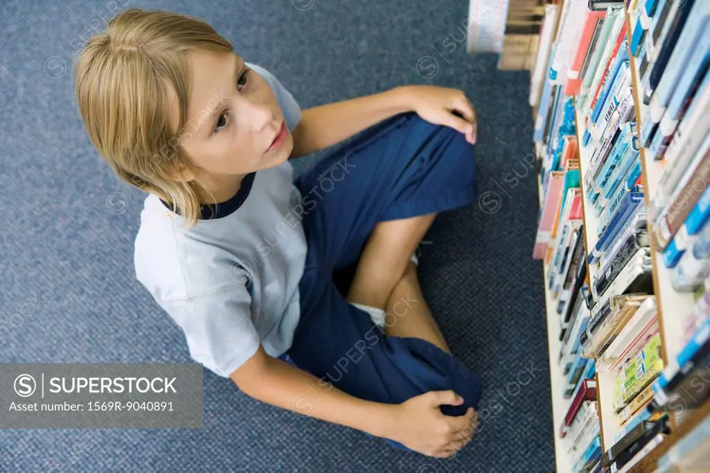 Boy sitting on floor looking up at rows of books lining bookcase