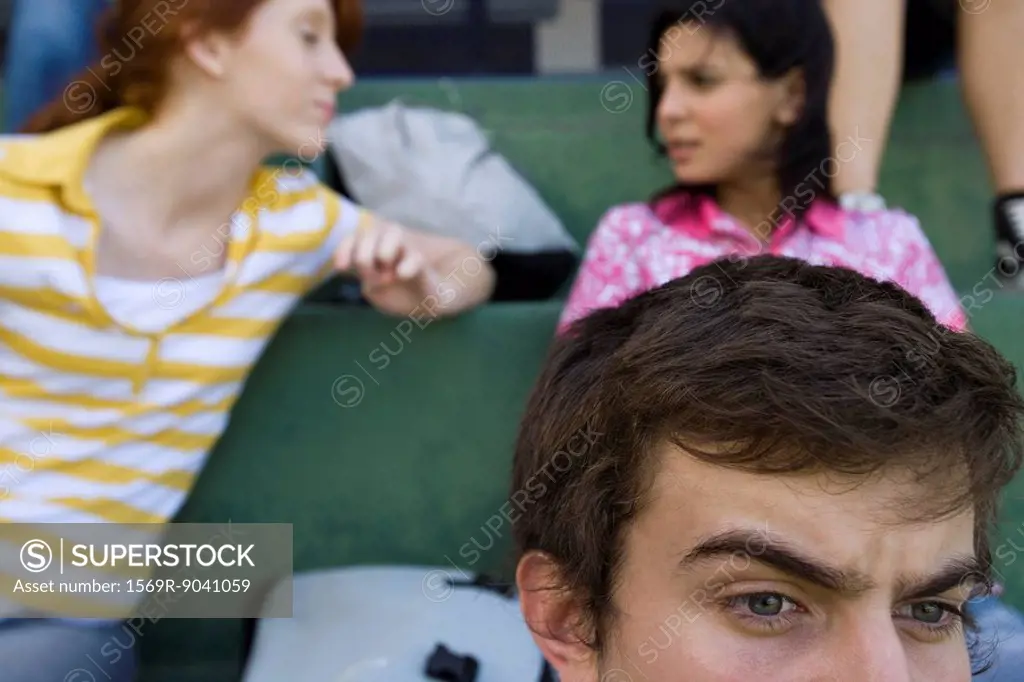 Hight school friends sitting together on bleachers, boy watching intently, girls in background chatting