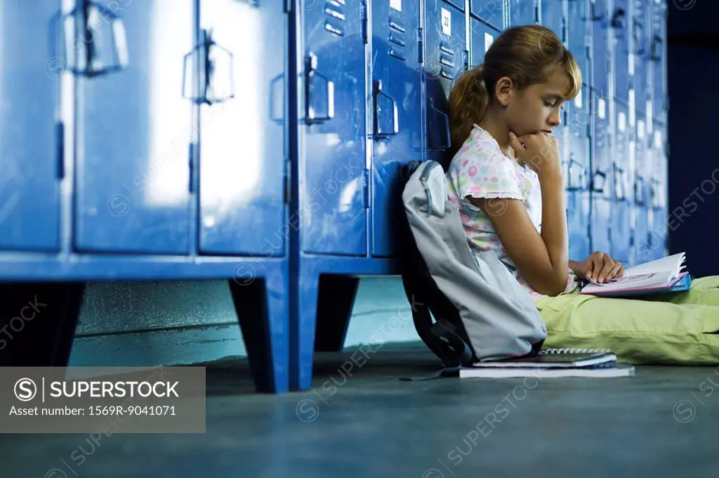 Female junior high student sitting on hall floor leaning against lockers reading book