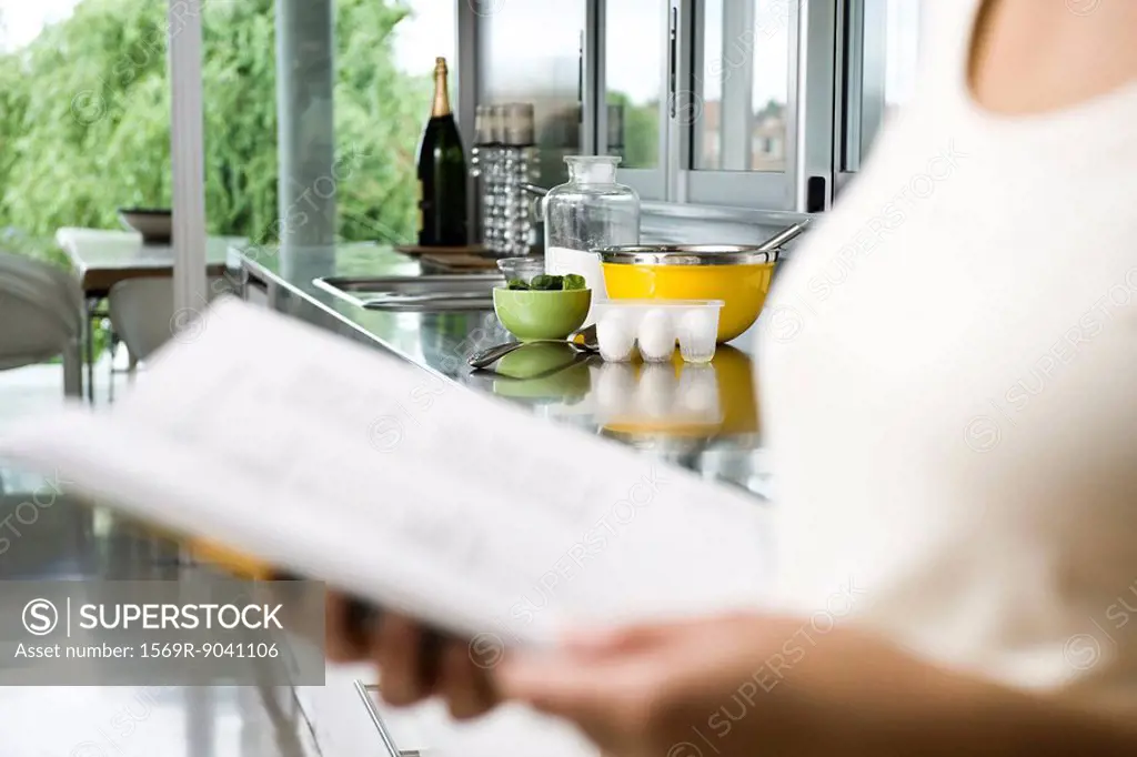 Reading recipe in cookbook, ingredients on kitchen counter in background