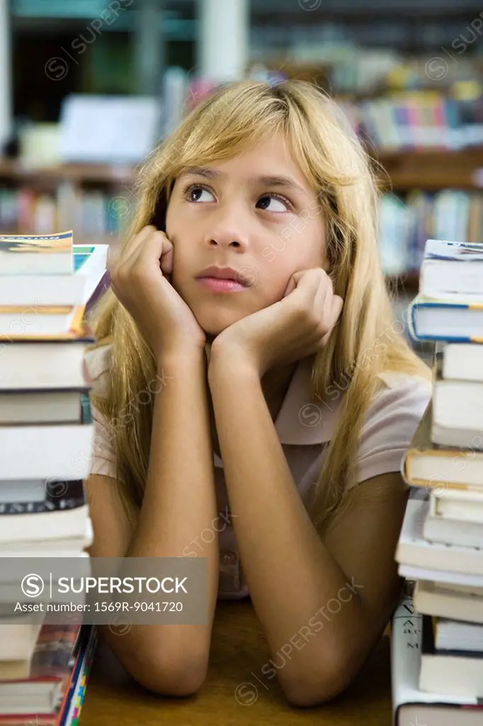 Preteen girl sitting with two large stacks of books, chin in hands, looking away