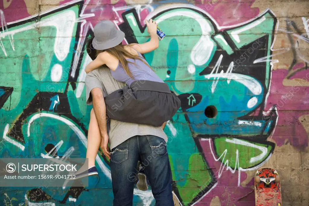 Young woman straddling companions back, spray painting graffiti mural