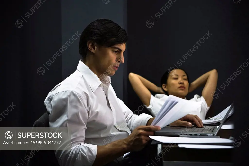 Man in office working late, colleague relaxing in background