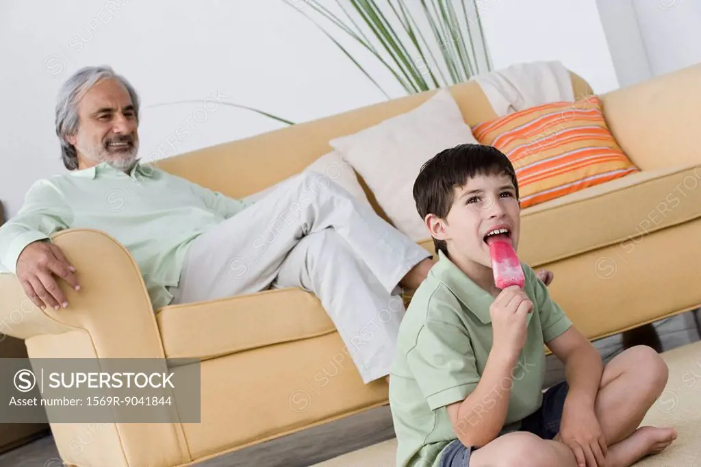Boy eating popsicle, grandfather relaxing in background