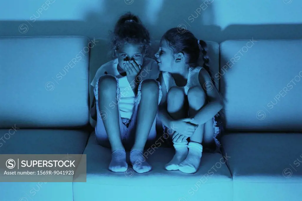 Two girls watching TV together, one whispering in the other´s ear
