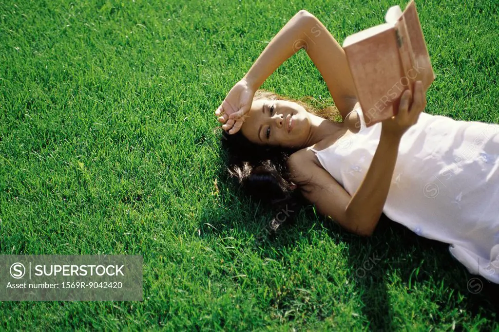 Young woman lying on grass, holding book, smiling at camera