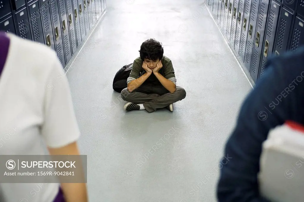 Teenage boy sitting sullenly on floor of school corridor, classmates in foreground