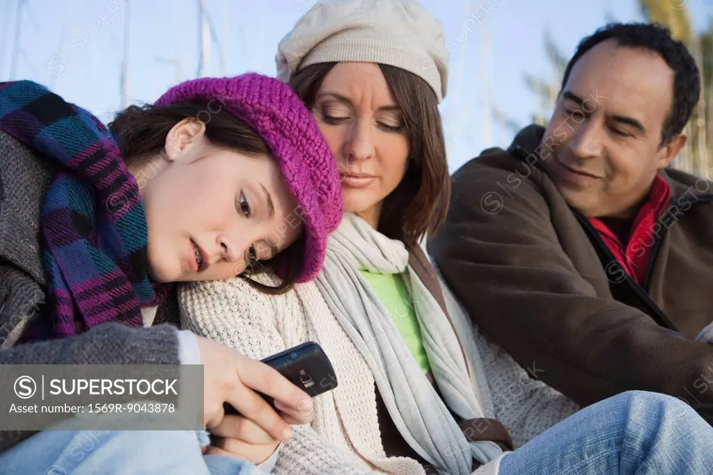 Teenage girl sitting outdoors with parents, text messaging with her cell phone