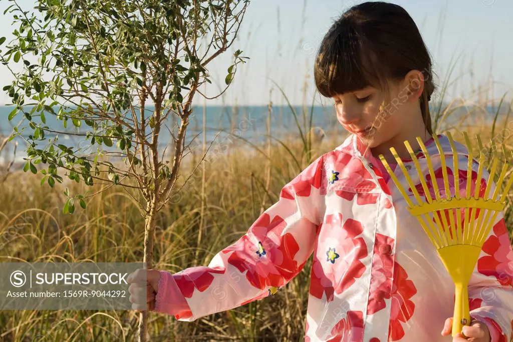 Girl with gardening rake touching trunk of sapling