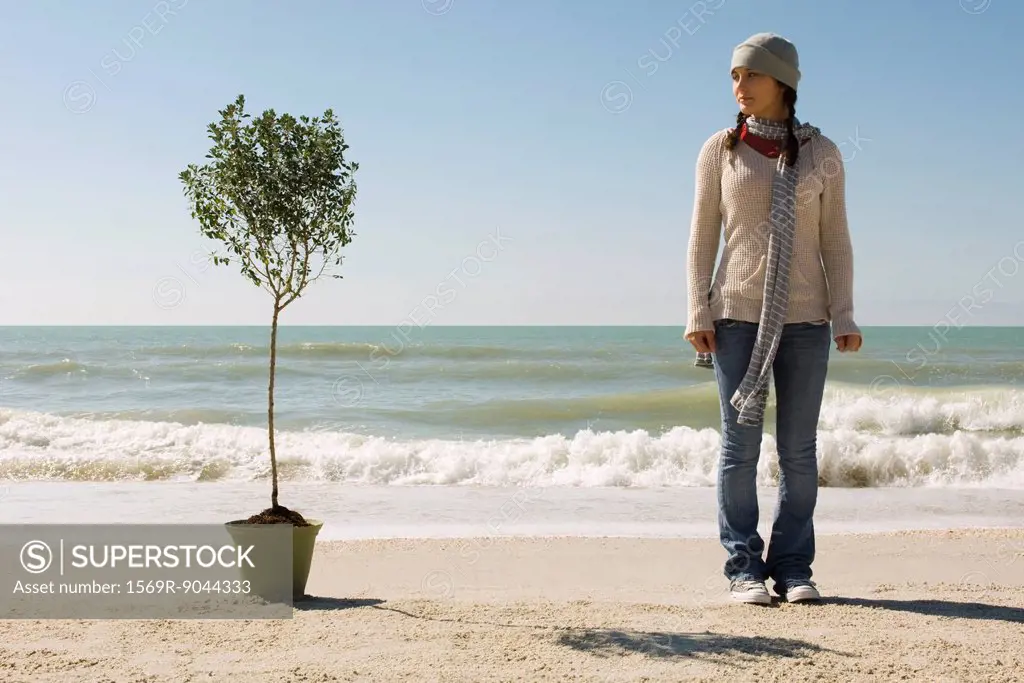 Preteen girl on beach standing near potted tree set near water´s edge