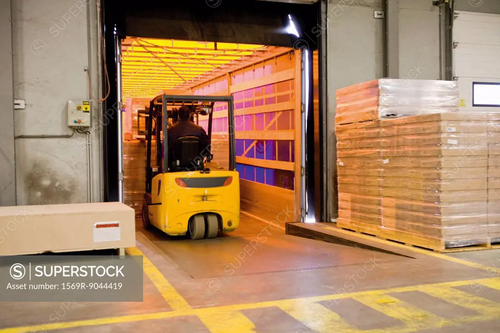 Forklift operator loading wrapped pallets of cardboard boxes onto trailer in warehouse