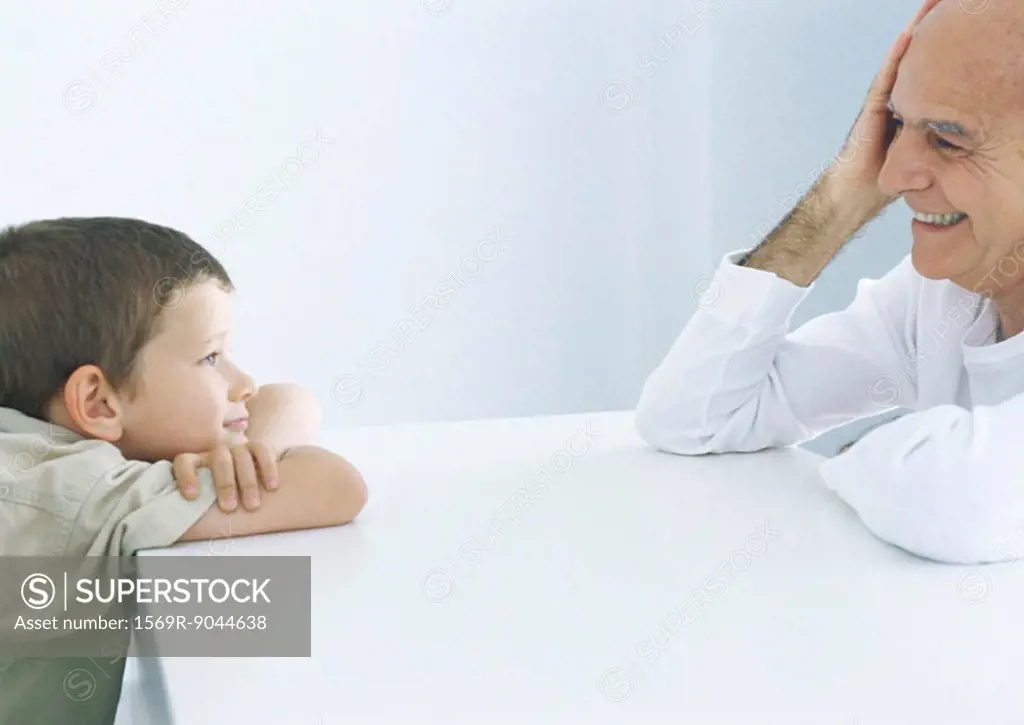 Boy and grandfather sitting across table, smiling at each other