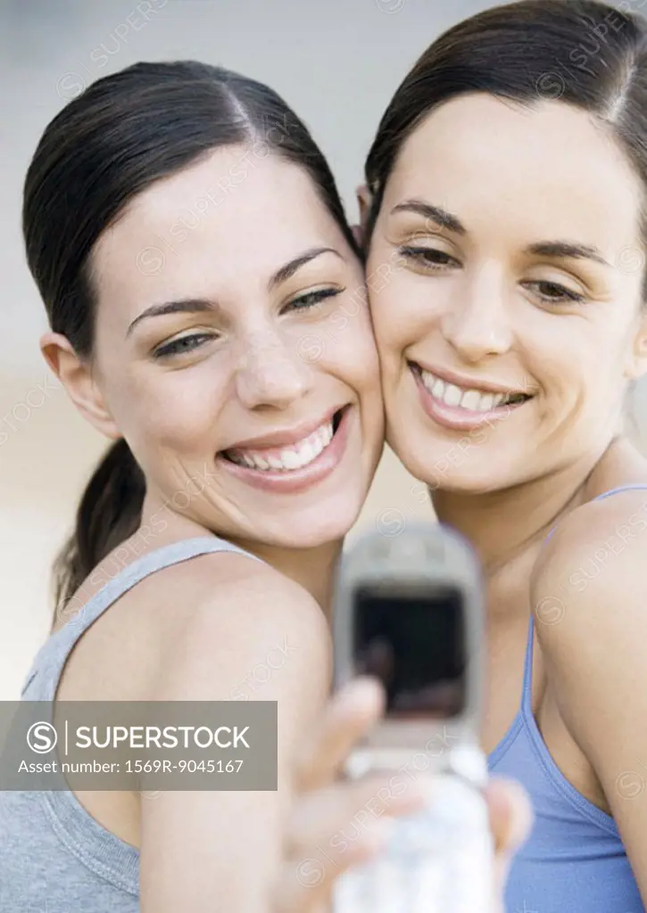 Two young female friends, cheek to cheek, taking photo with cell phone