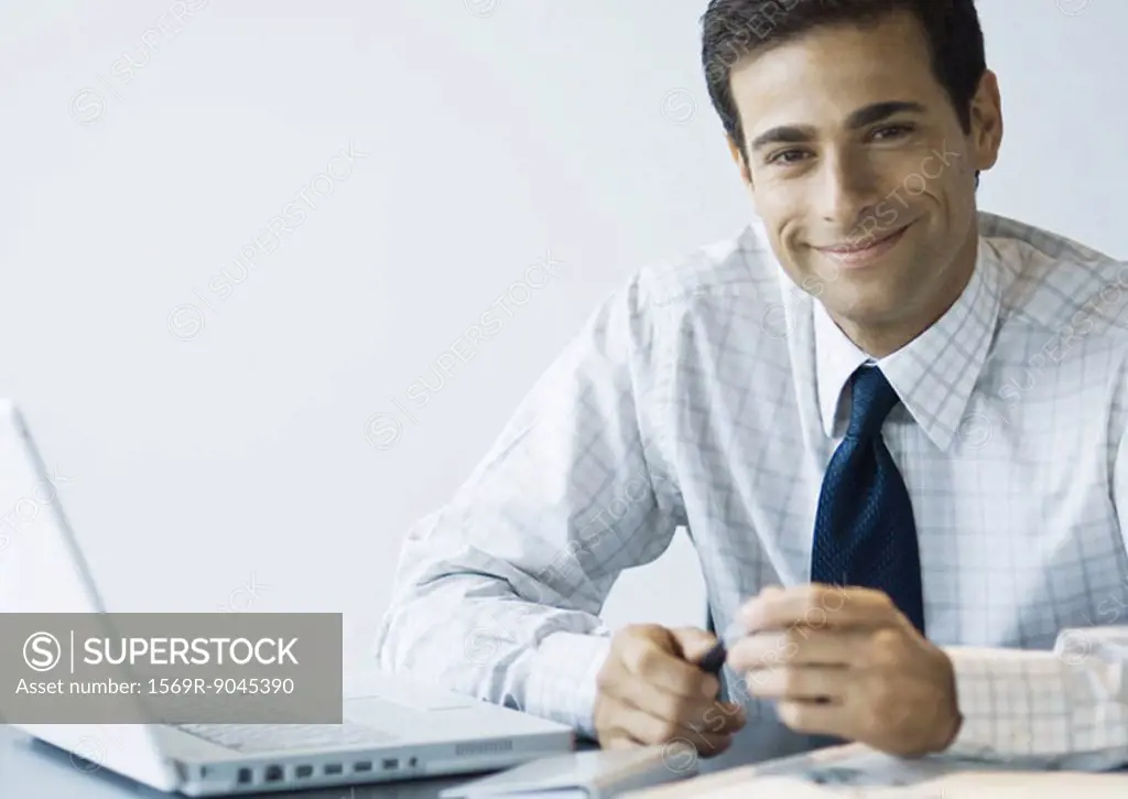 Businessman sitting at desk next to laptop, smiling at camera