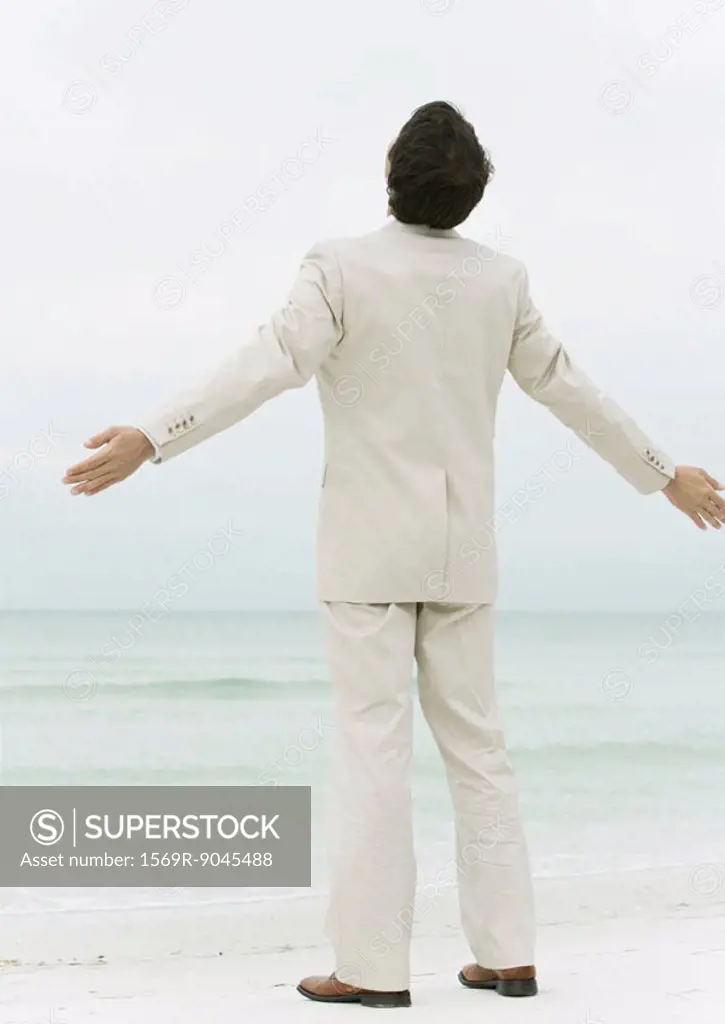 Businessman standing on beach with arms out, rear view