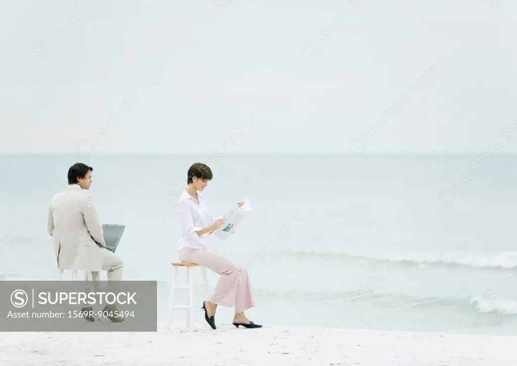 Businesspeople sitting on stools on beach, one using laptop, another reading newspaper