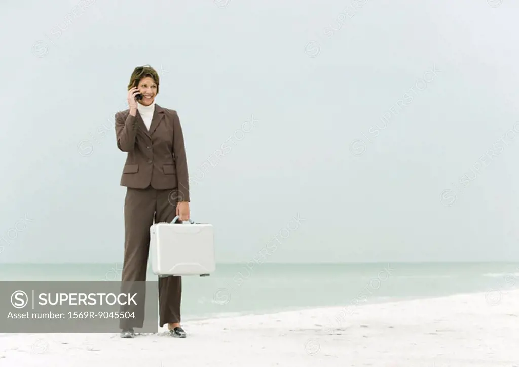 Businesswoman standing on beach holding briefcase, using cell phone