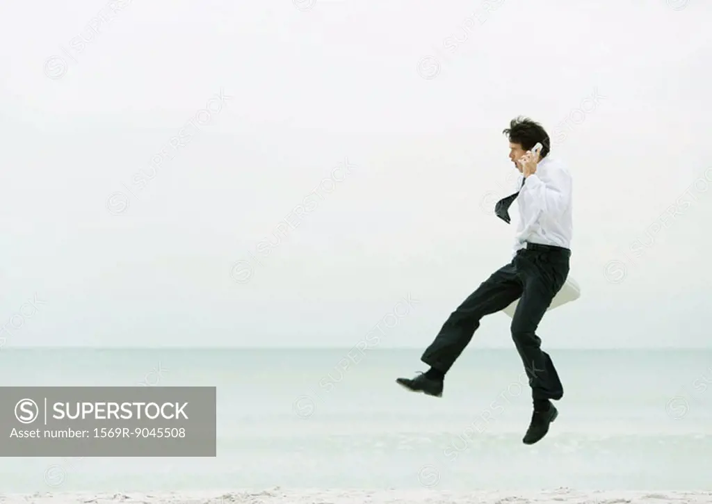 Businessman on beach, using cell phone and jumping