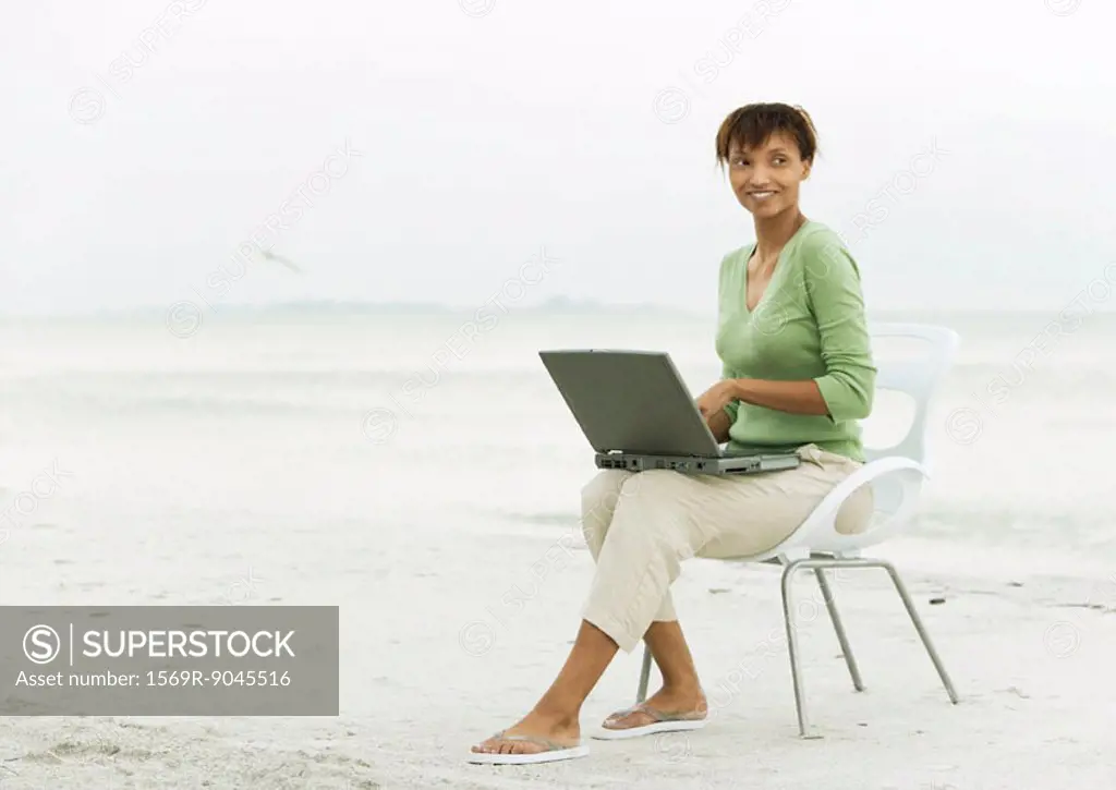 Woman on beach, sitting in chair using laptop