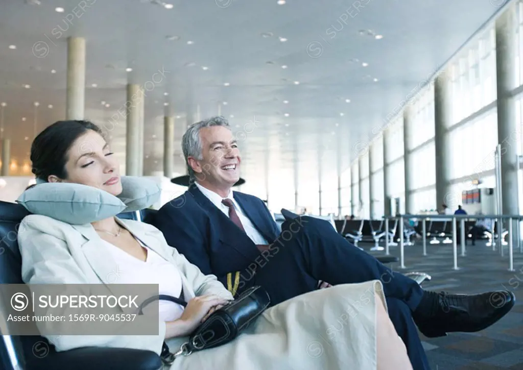 Business travelers sitting in airport lounge, woman napping with neck pillow next to smiling man