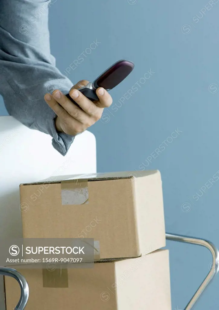 Man´s hand holding cell phone, cardboard boxes piled on chair