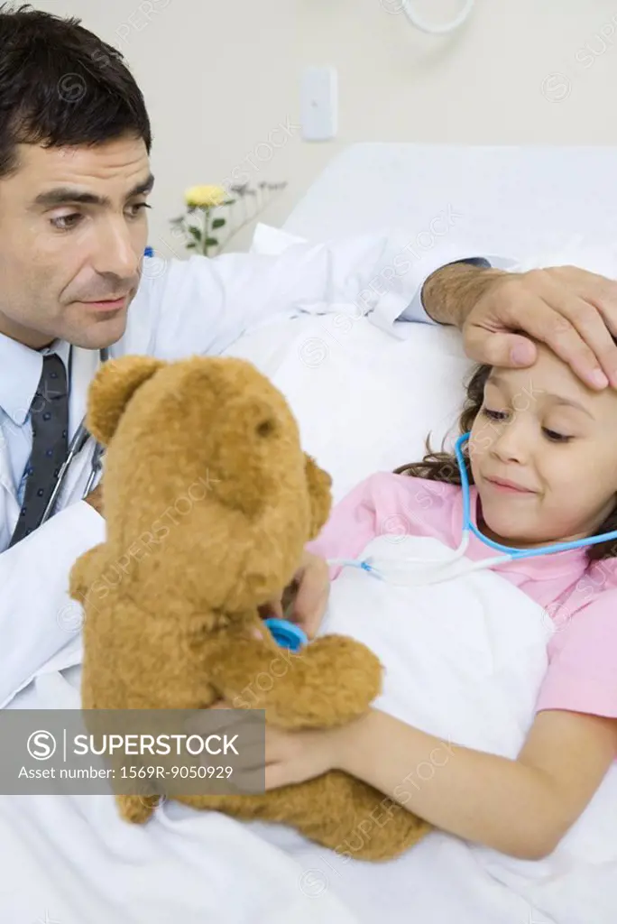 Child holding toy stethoscope to teddy bear, doctor feeling child´s forehead