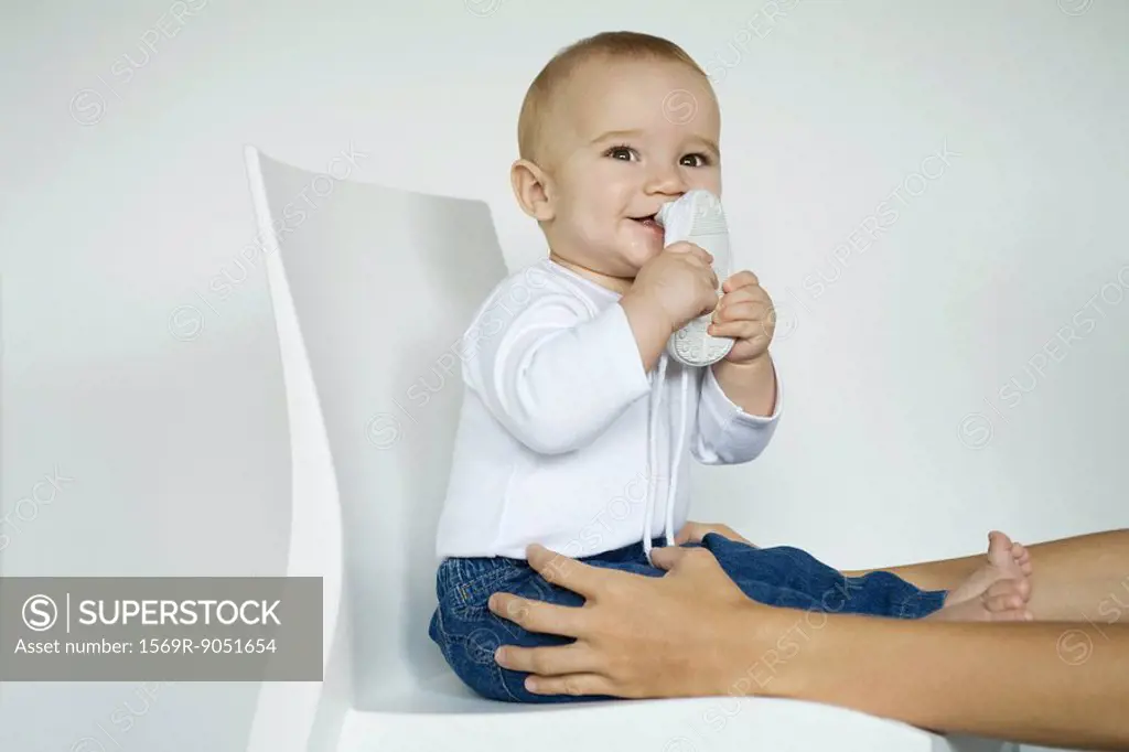 Baby sitting in chair putting shoe in mouth, mother´s arms holding baby´s legs