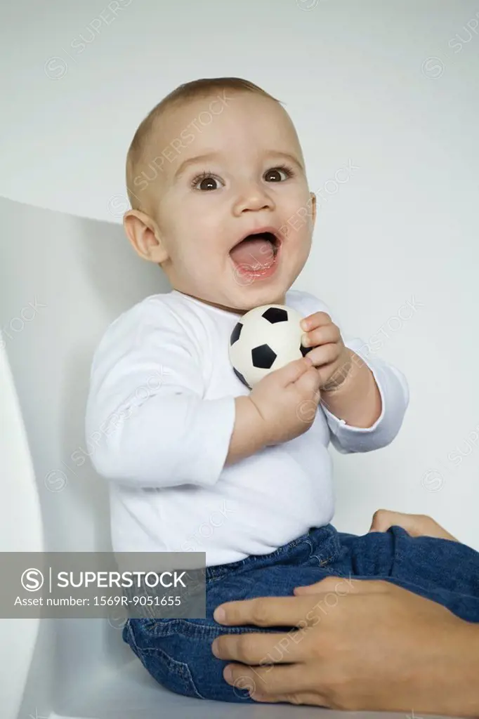 Baby sitting in chair holding ball, mother´s hands holding baby´s legs