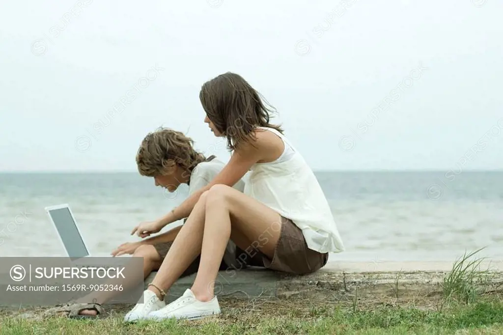 Teen girl and little brother sitting on the ground, using laptop computer, one pointing and looking over the other´s shoulder