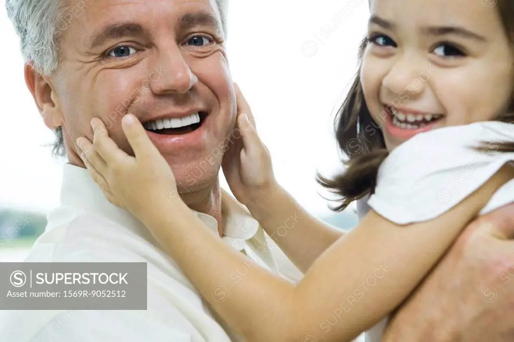 Man holding up little girl, girl´s hands on man´s cheeks, both smiling at camera, close-up