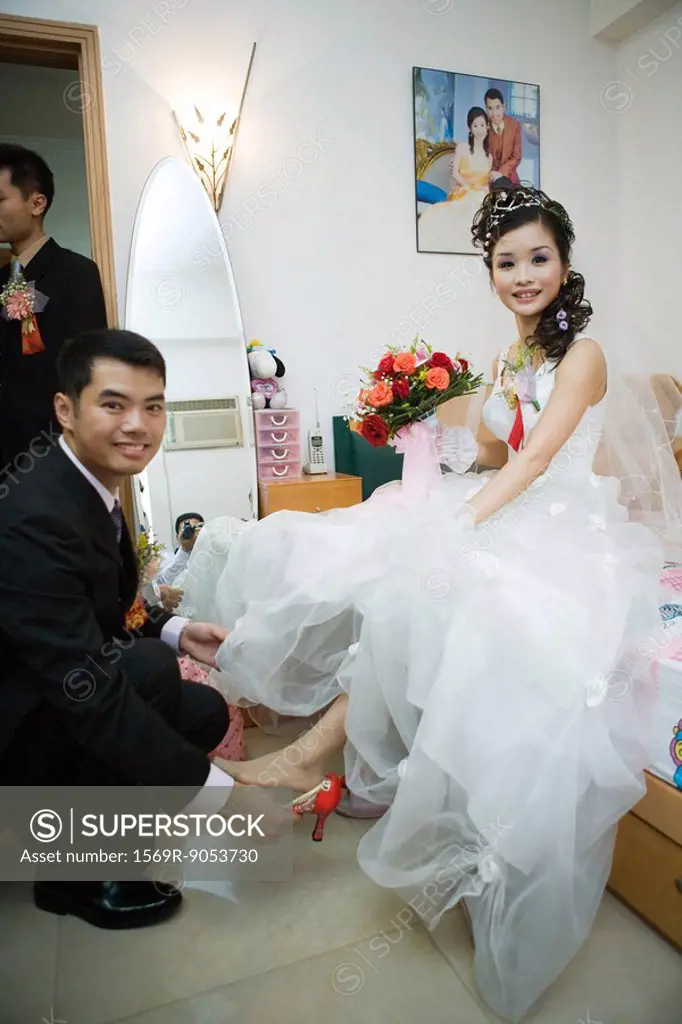 Groom putting shoe on bride´s foot in bedroom, both smiling at camera, full length