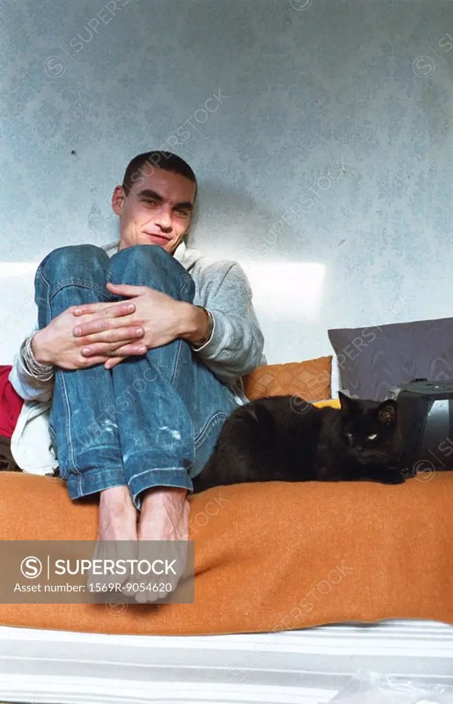 Young man sitting in bed hugging knees, cat lying alongside