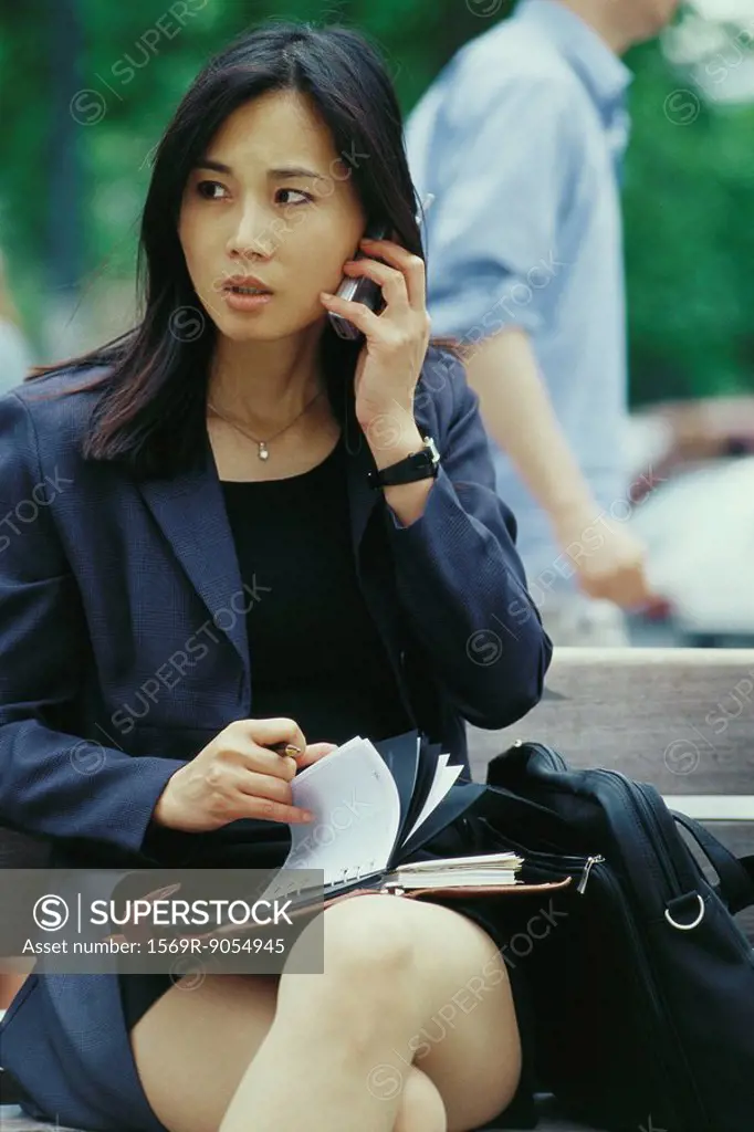 Businesswoman sitting on bench, using cell phone and flipping through agenda