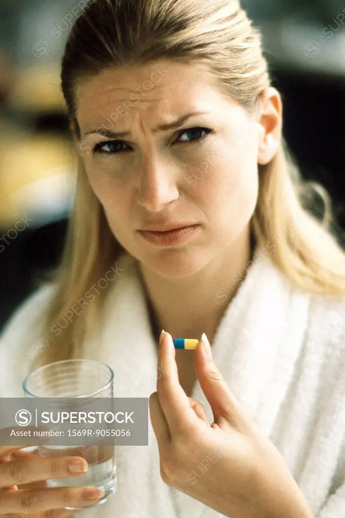 Woman holding pill and glass of water, frowning at camera