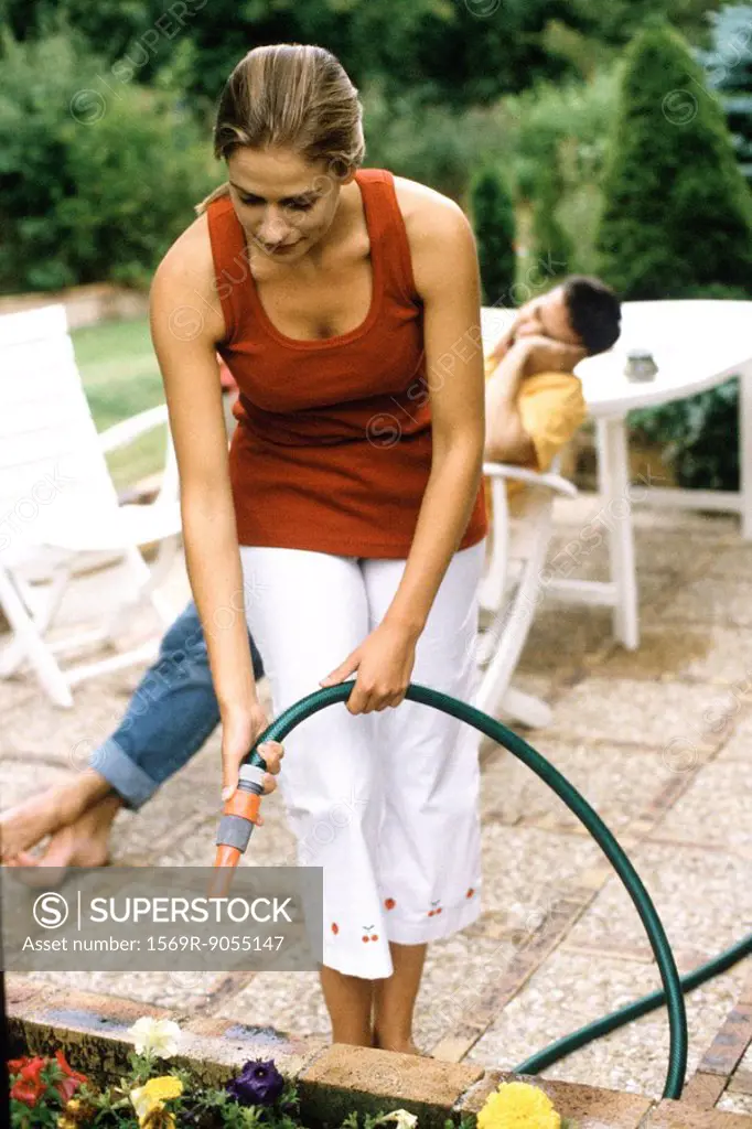 Woman watering flowers with garden hose, man lounging in background