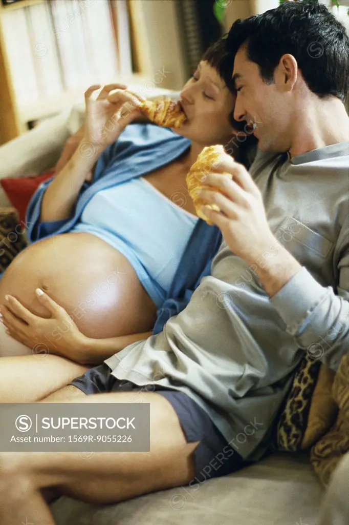 Expecting couple sitting together on sofa, eating croissants