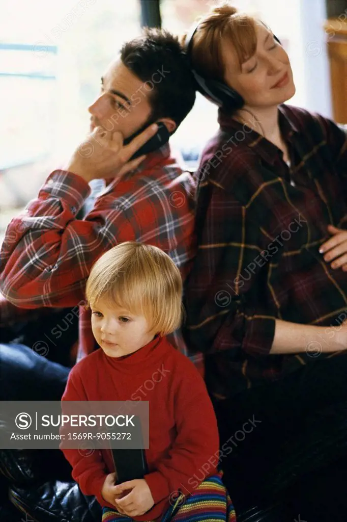 Family sitting back to back, father using cell phone, mother listening to headphones, daughter watching TV