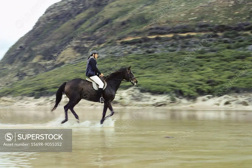 Woman riding horse in shallow water near rocky shore