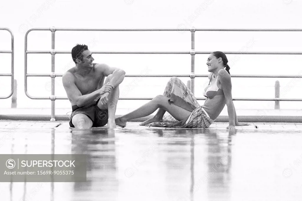 Young couple sitting together at poolside smiling at eachother