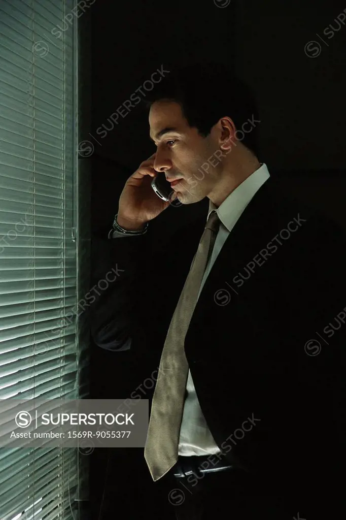 Businessman using cell phone, looking through blinds out window