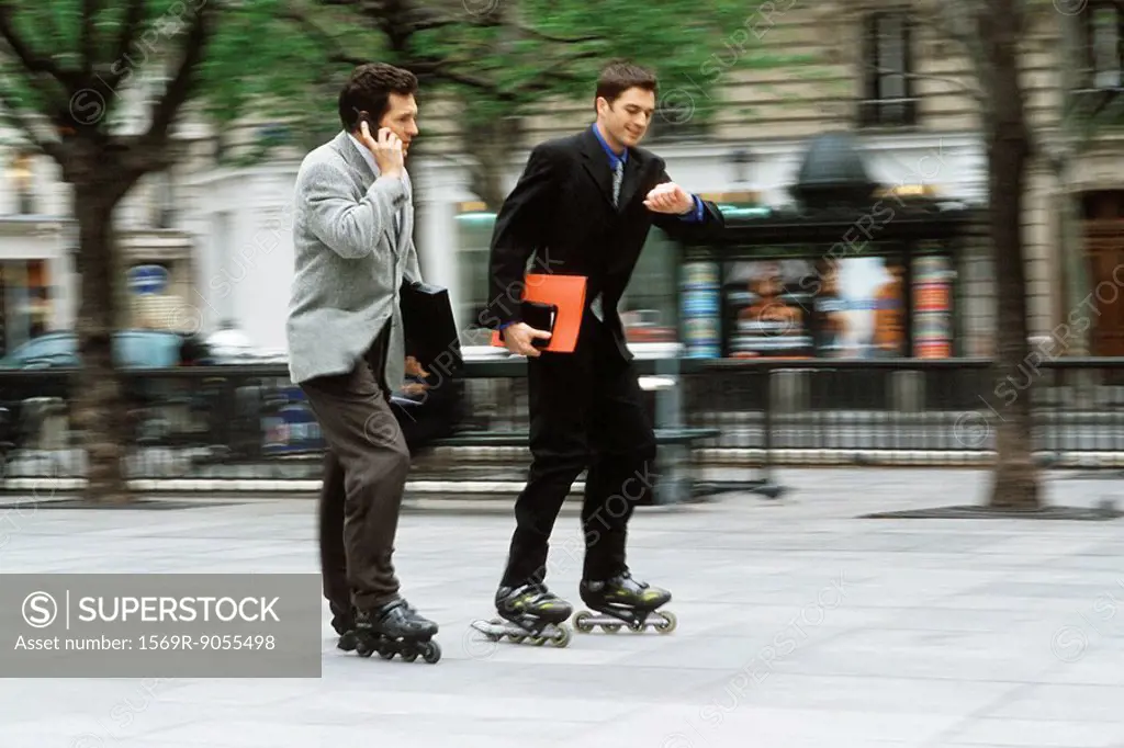 Men in business attire rollerskating together along sidewalk, one phoning, the other looking at watch
