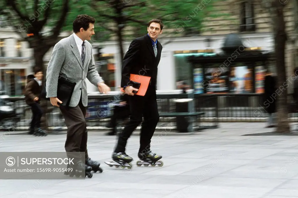 Men in business attire inline skating together along sidewalk carrying briefcase, folders under arm